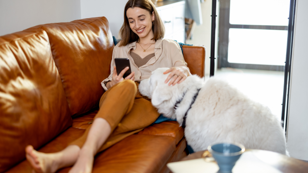 woman sits on couch looking at phone with large white dog beside her. She is petting the dog. 