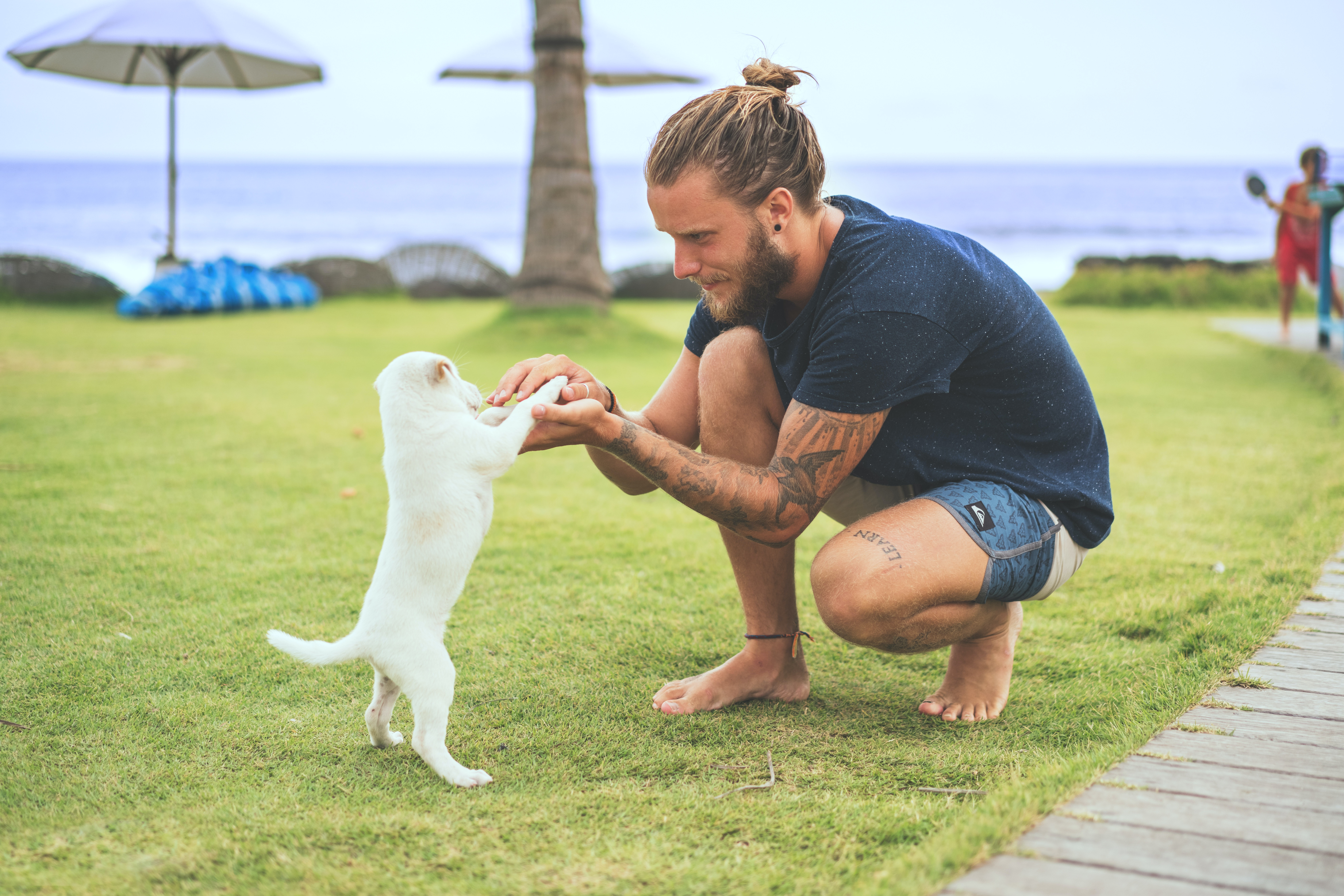 Man with beard and bun in navy tee shirt holds up small white puppy by the beach